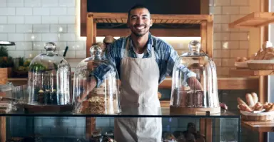 Dono de padaria sorrindo usando avental atrás do balcão do seu negócio, representando soluções de financiamento e acesso a crédito do NAC.